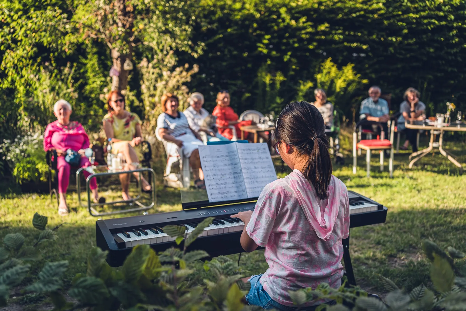 Ein Mädchen spielt im Garten Klavier, viele Menschen hören im Halbkreis im Garten zu.
